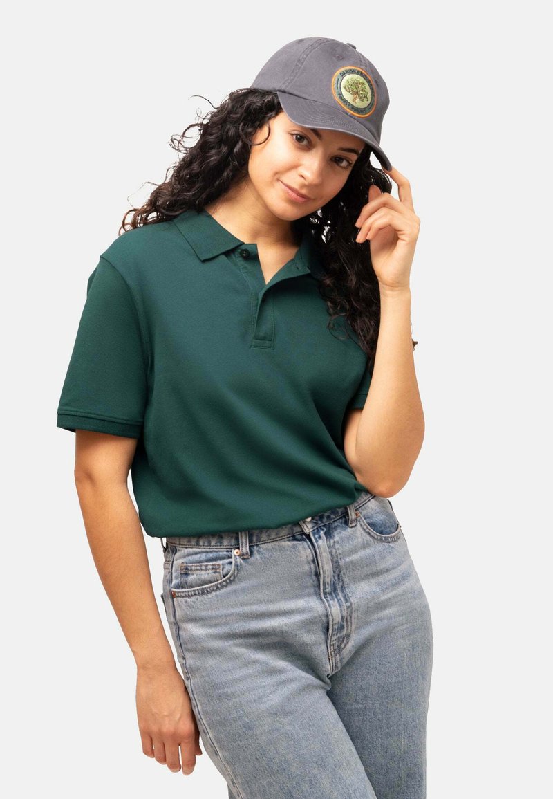 Polo in cotone verde scuro con una texture sottile, abbinato a jeans in denim blu chiaro. Un cappellino grigio presenta una toppa con logo circolare.