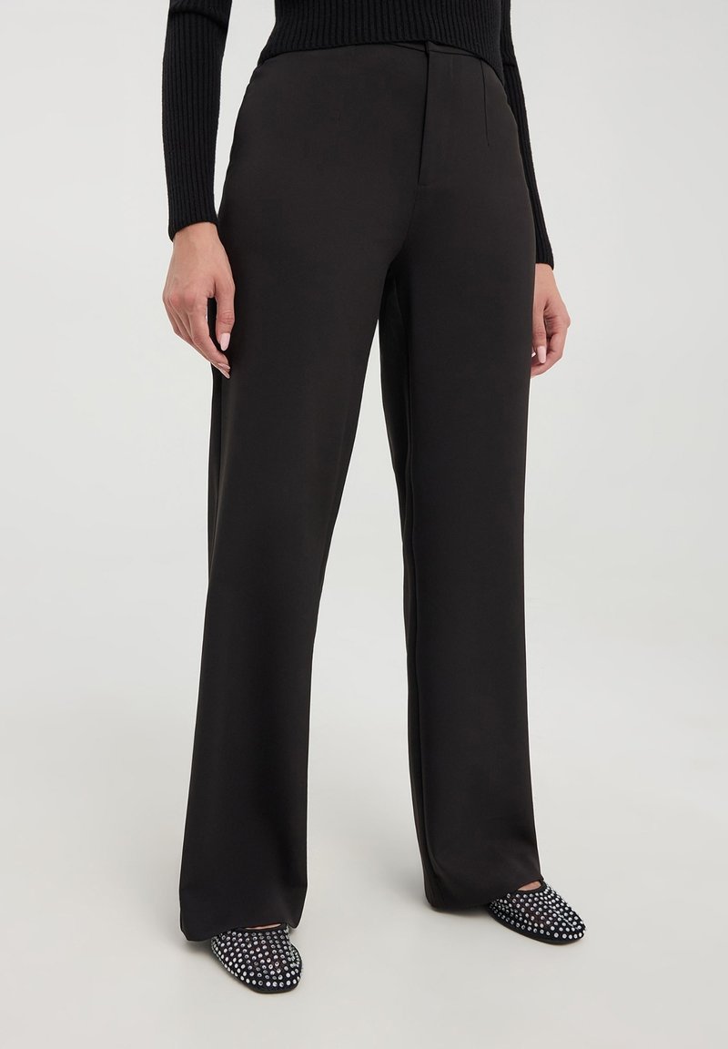Pantaloni neri su misura con un design a gamba dritta. Realizzati in tessuto liscio, presentano pince frontali e abbinati a mule a pois.