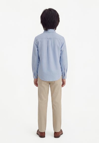 Kind staat met de rug naar voren, draagt een lichtblauw shirt met lange mouwen, beige broek en bruine nette schoenen tegen een effen witte achtergrond.