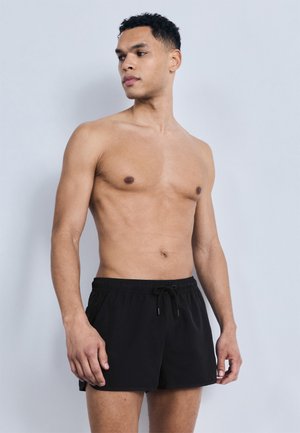 Muž bez košele v čiernych plavkách so sťahovacou šnúrkou, stojaci pred jednoduchým svetlošedým pozadím, pozerajúci sa do strany.