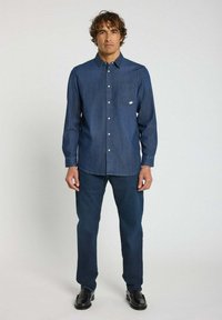 Chemise en denim bleu foncé avec col boutonné, manches longues et poche poitrine. Assortie avec un pantalon bleu foncé et des chaussures noires.