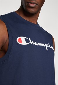 Mornarska majica bez rukava od pamuka s okruglim izrezom, s velikim bijelim 'Champion' logom i crvenim naglaskom na slovu 'C'.