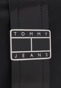 Sac à dos en nylon noir avec un logo rectangulaire argenté brillant affichant "TOMMY JEANS" en lettres majuscules sur un fond noir.