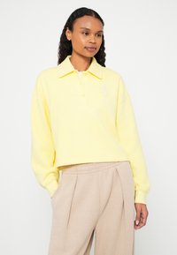 Camisola amarela curta com gola, apresenta uma pala com três botões e um pequeno detalhe de logótipo. Textura macia, ajuste descontraído e mangas compridas.