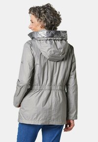 Lichtgrijze waterdichte jas met een capuchon, elastische taille, glanzende zilveren kraagaccent en een zachte textuur. Beschikt over lange mouwen en een losse fit.