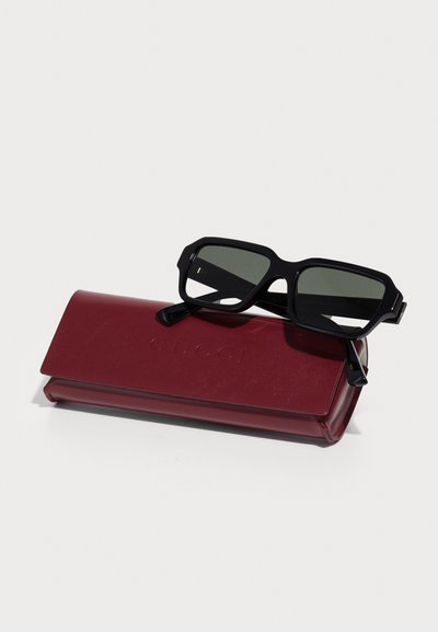 Lunettes de soleil rectangulaires noires avec des verres foncés, reposant sur un étui rigide bordeaux embossé avec "Gucci." L'étui présente une texture lisse.