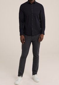 Muž oblečený v černé košili s knoflíky, tmavě šedých slim-fit kalhotách a bílých teniskách stojí proti neutrálnímu pozadí.