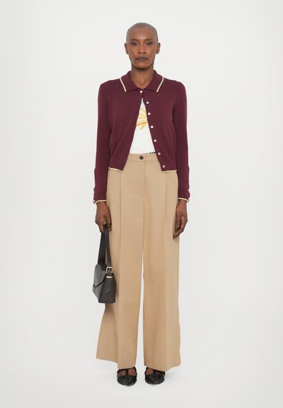 Cardigan bordeaux avec des détails jaunes, t-shirt graphique blanc, pantalons larges beiges, sac à bandoulière noir et ballerines tressées noires.
