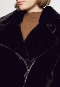 Sort jakke i imiteret pels med bredt revers, sølv lynlåsdetalje og blød tekstur, kombineret med en brun ribstrikket sweater under.