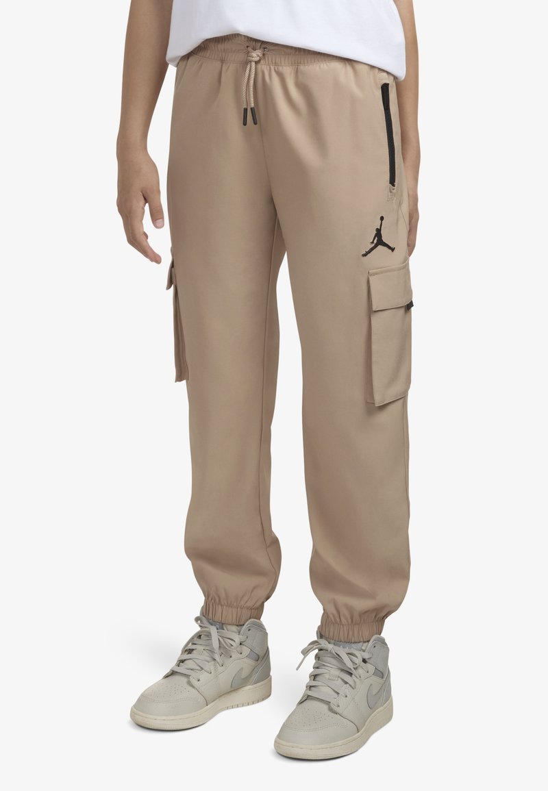 Pantalon jogger cargo beige avec poignets élastiques, poches latérales à rabat, une poche zippée noire et un logo Jumpman noir sur la cuisse droite.