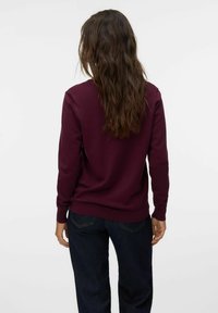 Pullover bordeaux con maniche lunghe, scollo a giro e polsini a costine. Indossato sopra jeans di denim scuro, mostra una vestibilità comoda.