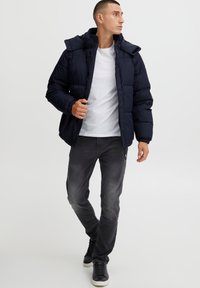 Mørkeblå dunjakke med hette, glidelåslukking og elastiske mansjetter, kombinert med grå jeans og svarte sneakers. Jevnt materiale og quiltet design.