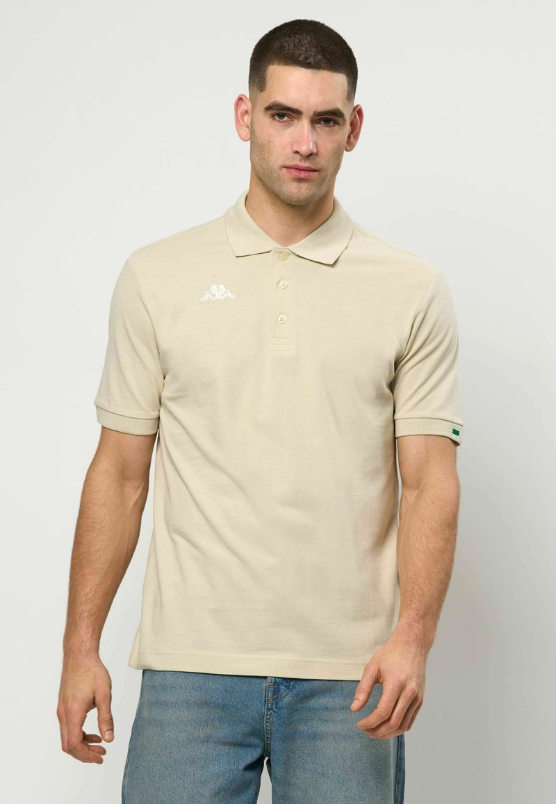 Homme aux cheveux courts portant un polo beige avec un logo blanc sur la poitrine et un jean bleu, debout devant un fond uni.
