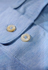 Jasnoniebieska koszula materiałowa o gładkiej fakturze, z dwoma kremowymi guzikami przyszytymi niebieską nitką w pobliżu kołnierzyka.