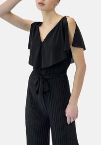 Svart jumpsuit med en volangfärgad, lager på lager-överdel, v-ringning och knytning i midjan. Nertill har den en veckad struktur för ökad visuell intress.