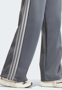 Sivé športové nohavice z hladkého materiálu s tromi vertikálnymi bielymi pruhmi pozdĺž strán, skombinované so svetlými teniskami.