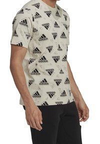 Mies, joka pukeutuu vaaleanbeigeen t-paitaan, jossa on toistuvia mustia Adidas-logoja ja geometrisia muotoja, yhdistettynä mustiin housuihin.