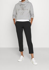 Hoodie grigio corto con la scritta "TRUE RELIGION 2002" in lettere metalliche, abbinato a pantaloni neri a sigaretta e sneakers bianche.