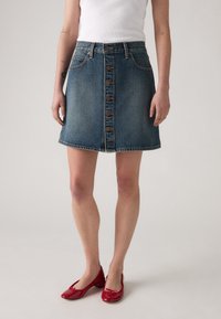 Falda de mezclilla en corte A, lavado azul claro, cierre frontal con botones, dos bolsillos laterales, dobladillo con bordes deshilachados, combinada con zapatos de charol rojos.