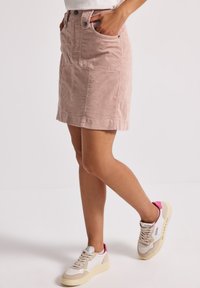 Kvinna som bär en knälång dammig rosa kordflorskjol och vita sneakers med rosa detaljer, ståendes med ett ben lätt böjt.