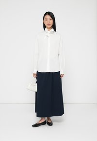 Camicia bianca a maniche lunghe con colletto a farfalla, abbinata a una gonna maxi plissettata blu navy. Porta una piccola borsa bianca arricciata. Scarpe ballerine nere.