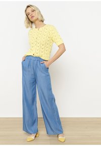 Rumena pletena kratka majica s kratkimi rokavi in gumbi spredaj, kombinirana z ohlapnimi širokimi modrimi jeans hlačami in rumenimi čevlji na blok peto.