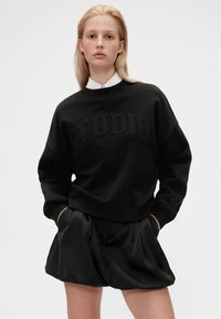 Femme blonde portant un sweatshirt noir avec le texte "STUDIO" et un short volumineux noir, se tenant les mains dans les poches contre un fond uni.
