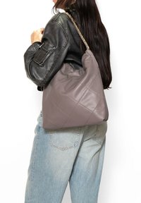 Borsa a tracolla in pelle trapuntata grigia con forma morbida, dotata di una tracolla a catena. Indossata sulla spalla, abbinata a una giacca di pelle nera e jeans chiari.