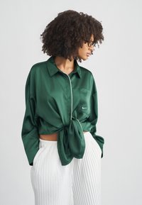 Πράσινο σατέν πουκάμισο με δέσιμο μπροστά, με γιακά και τσέπη στο στήθος. Συνδυασμένο με λευκό πλισέ παντελόνι.