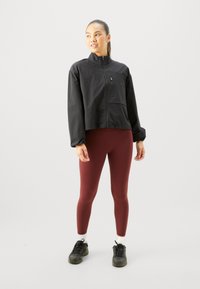 Schwarze Cropped-Jacke mit hohem Kragen, strukturiertem Stoff und einer vorderen Reißverschlusstasche, kombiniert mit bordeauxfarbigen Leggings und schwarzen Sneakers.