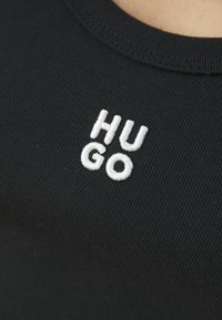 Juodas įgulos tipo marškinėliai su balta siuvinėta tekstu "HUGO" priekyje. Apvalus iškirpimas ir prigludusi konstrukcija.
