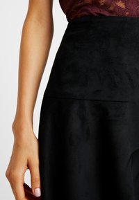 Falda de ante negro con una textura suave, presenta una cintura alta y sin patrones visibles. Una mano está colocada en la costura lateral.