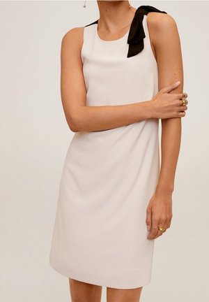 Beżowa, bezrękawna sukienka z czarną kokardą na ramieniu, noszona przez osobę z złotymi pierścionkami na palcach i skrzyżowanymi ramionami na piersi.