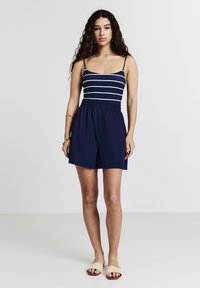Rompers blu navy con strisce orizzontali bianche, spalline regolabili e vita elastica. Abbinati a sandali scivolabili in paglia beige.