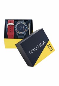 Nautica laikrodis su juodu ciferblatu, daugiaspalviais akcentais, dviem silikoniniais dirželiais (raudonu ir mėlynu) bei įrankiu dirželiui keisti, pateiktas geltonoje ir tamsiai mėlynoje dėžutėje.