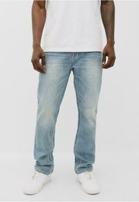 Vaqueros de denim azul claro con corte recto, que presentan un desgastado sutil, cinco bolsillos y un cierre de botones. Combinados con zapatillas blancas.