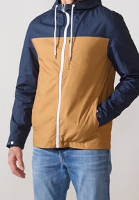 Färgblockerad jacka med marinblå övre del och bruna nedre sektioner, med en vit dragkedja, huva med dragsko och två framfickor.