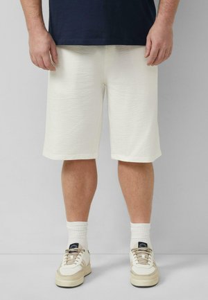 Mann trägt strukturierte off-white knielange Shorts, weiße Socken und beige Sneaker, steht vor einem einfarbigen grauen Hintergrund.