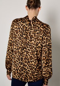 Bluzka w wzór panterki z gładkiego materiału, z wysokim dekoltem, wycięciem na plecach i długimi rękawami z mankietami zapinanymi na guziki. Ciemnobrązowe i jasnobrązowe odcienie.