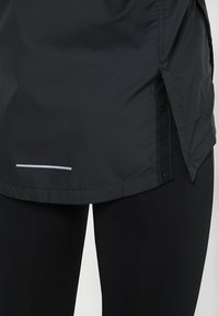 Veste imperméable noire avec une texture lisse, un ourlet courbé, des boutons sur le côté et une bande réfléchissante près du bas pour plus de visibilité.