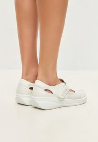 White, breathable sports shoes with a thick sole, featuring a Velcro strap and cut-out details on the sides for added ventilation.