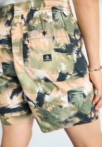 Shorts de camuflaj dintr-un material ușor, cu modele de pensule în verde și bej, un brâu elastic și un buzunar pe spate cu logo.