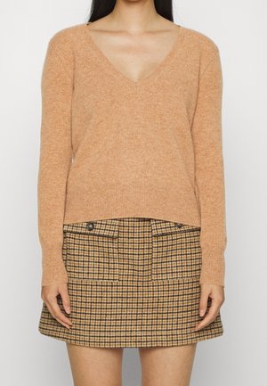 Vrouw die een camelkleurige trui met V-hals draagt, gecombineerd met een hooggetailleerde, bruin-zwart geruite minirok met knopen aan de voorkant en zakken.