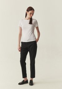 Haut blanc ajusté à manches courtes associé à un pantalon noir sur mesure et des chaussures slip-on noires avec des détails texturés, debout devant un fond clair.