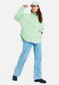 Kvinna i en ljusgrön oversized kordjacka, ljusblå jeans, svarta stövlar och vit basebollkeps, stående med en hand upplyft.