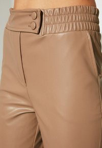 Hoch geschnittene, tanfarbene Kunstlederhose mit einem gerafften Elastikbund, ausgestattet mit zwei dekorativen Knöpfen und einer glatten, glänzenden Textur.