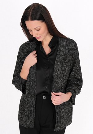 Frau mit langen dunklen Haaren, die ein schwarzes, geknöpftes Hemd und eine dunkelgraue gestrickte Strickjacke trägt, schaut nach unten, während sie die Strickjacke richtet.