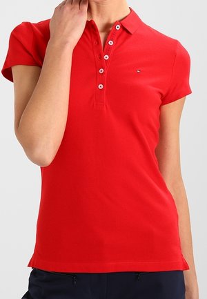 Frau trägt ein enganliegendes rotes Poloshirt mit kurzen Ärmeln, fünf Knöpfen und einem kleinen Logo auf der linken Brust.