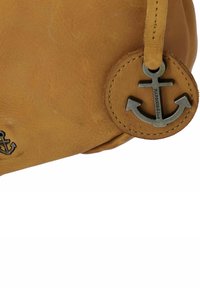 Angolo di una borsa in pelle marrone con un ciondolo a forma di ancora in metallo inciso "HARBOUR & 2nd" cucito su una targhetta rotonda in pelle.