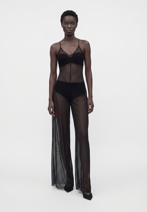Femme portant un body en maille noire transparente avec un soutien-gorge intégré et un sous-vêtement taille haute, debout devant un fond clair uni.